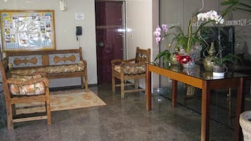Um hall de hotel com balcão de recepção, dois sofás de madeira, uma mesa de jantar e uma orquídea em vaso.