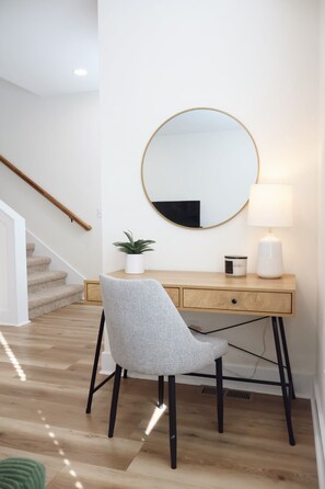 Una habitación moderna con un escritorio de madera, una silla gris, un espejo redondo, una planta en maceta y una lámpara.