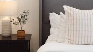Een keurig opgemaakt bed met witte beddengoed en gestreepte kussens, een bedlampje en een potplantje op een nachtkastje van donker hout.
