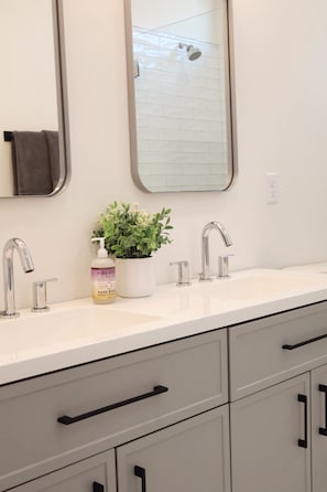 Une salle de bain avec un comptoir blanc, deux lavabos, un miroir, un porte-serviettes, une plante en pot et un flacon de savon pour les mains.