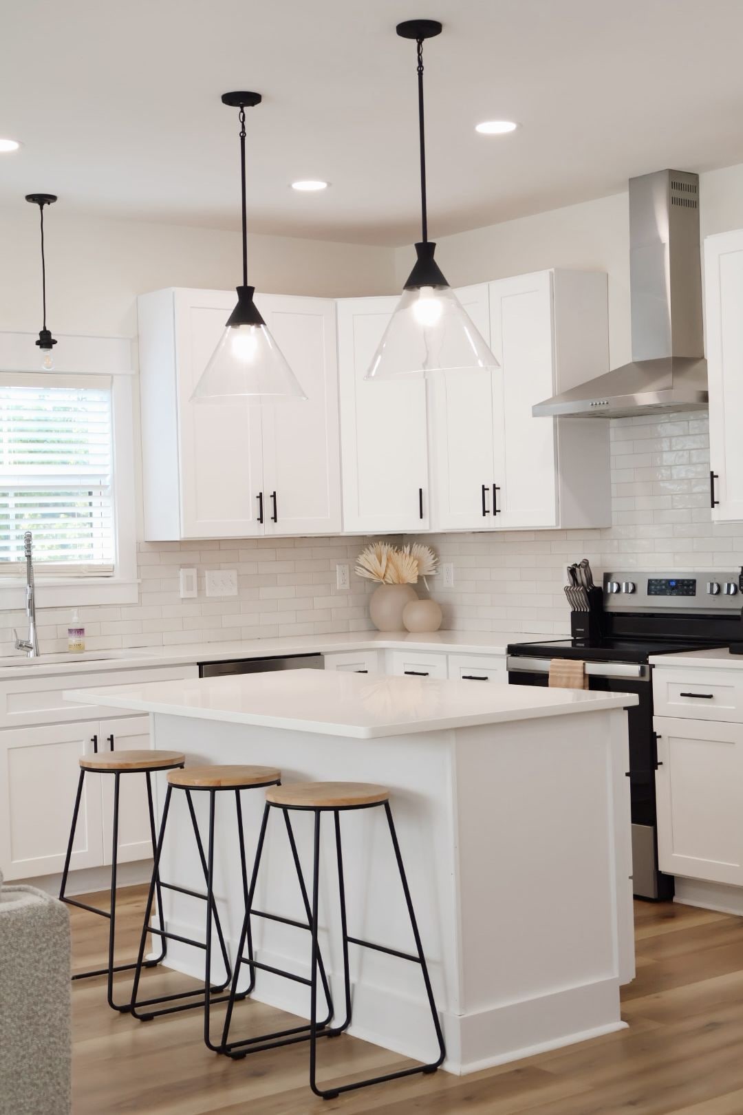 A modern kitchen with white cabinetry, a central island with stools, pendant lighting, and a stainless steel range hood.