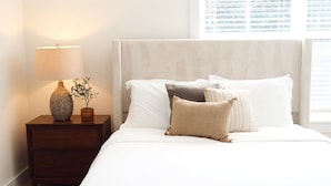 Een opgemaakt bed met witte lakens en meerdere kussens, een bedlampje en een kleine potplant op een houten nachtkastje.
