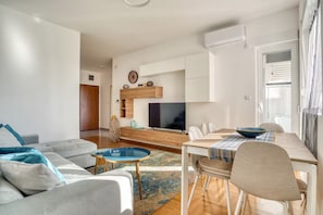 A modern living room with a sofa, coffee table, TV, and dining area with chairs and a table. - Baby Blue Apartment (Budva)
