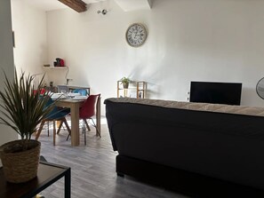 Un salon moderne comprenant un canapé, une table à manger avec des chaises, un petit bureau avec un ordinateur, une horloge murale et une plante en pot.