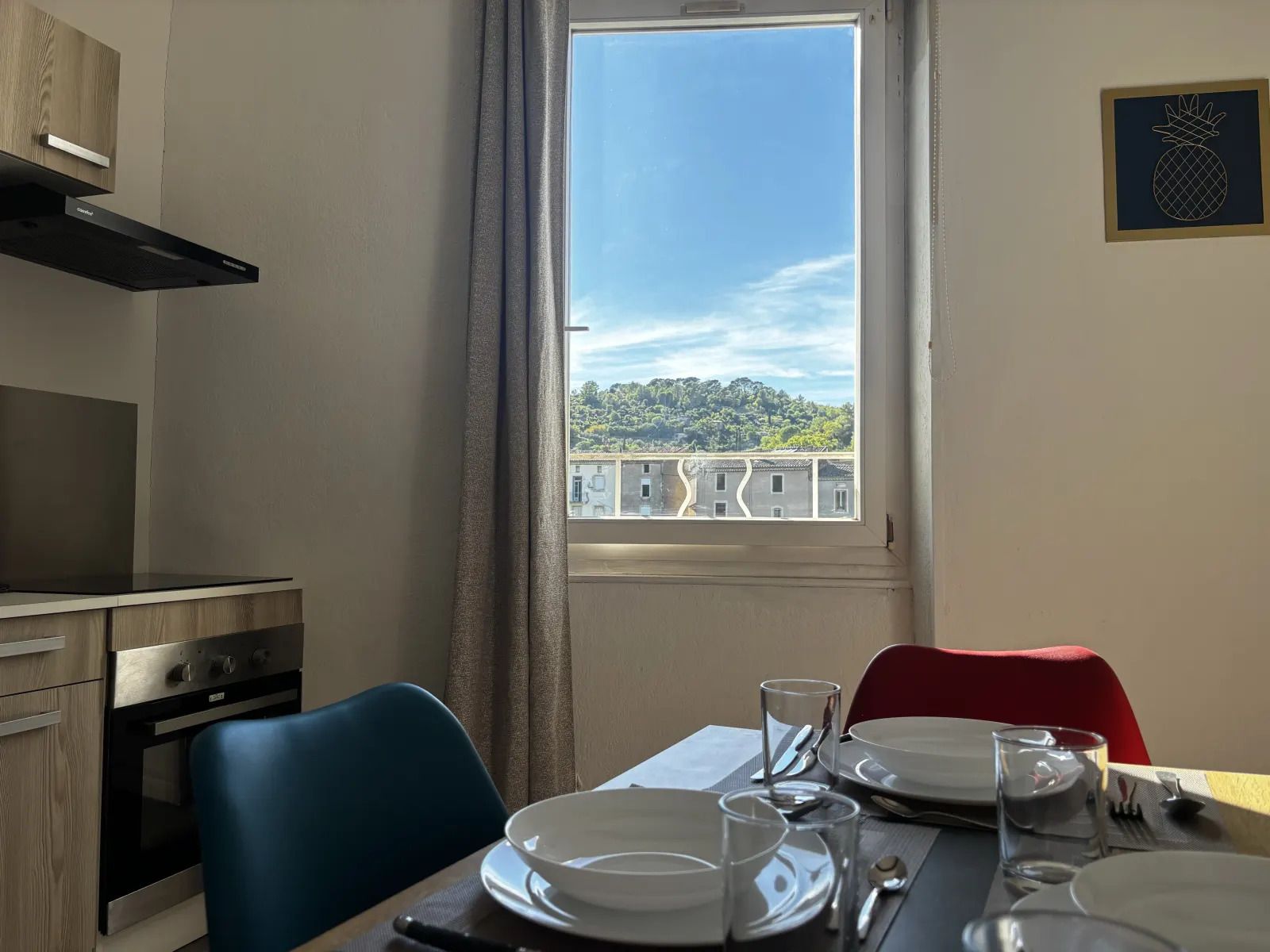 A dining area with a table set for two, a window with a view of buildings, and a kitchen with modern appliances.