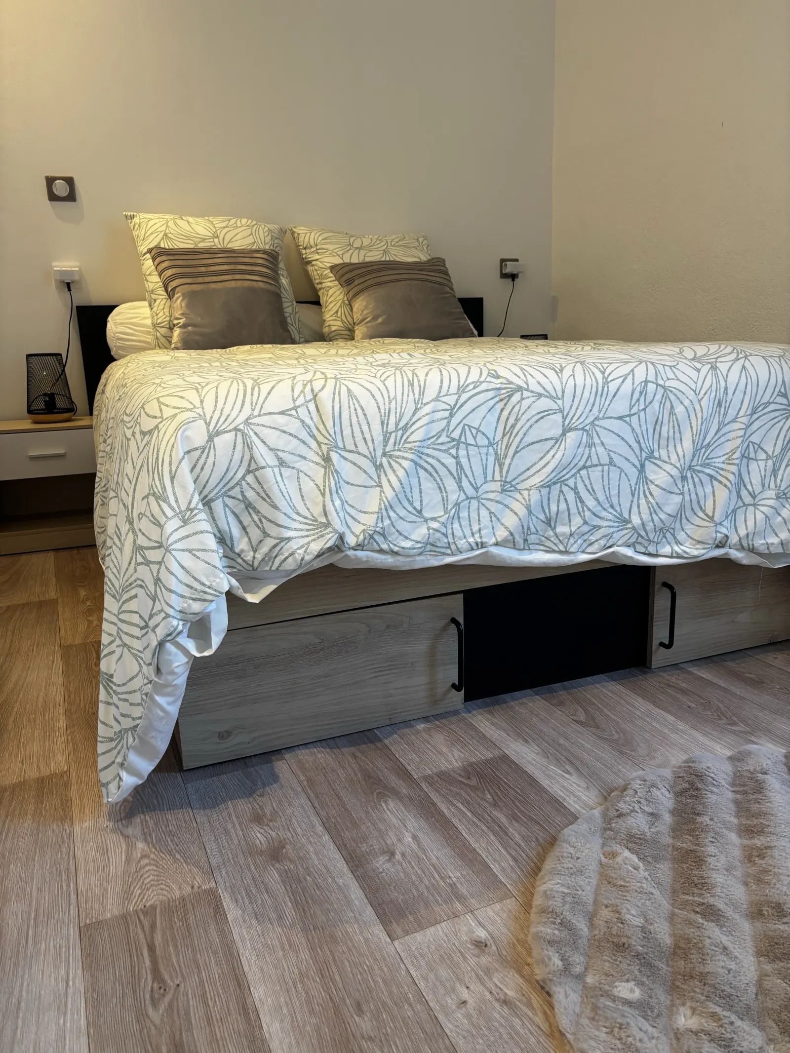 A bed with a leaf-patterned bedspread, a bedside table with a lamp, and a power outlet on the wall.