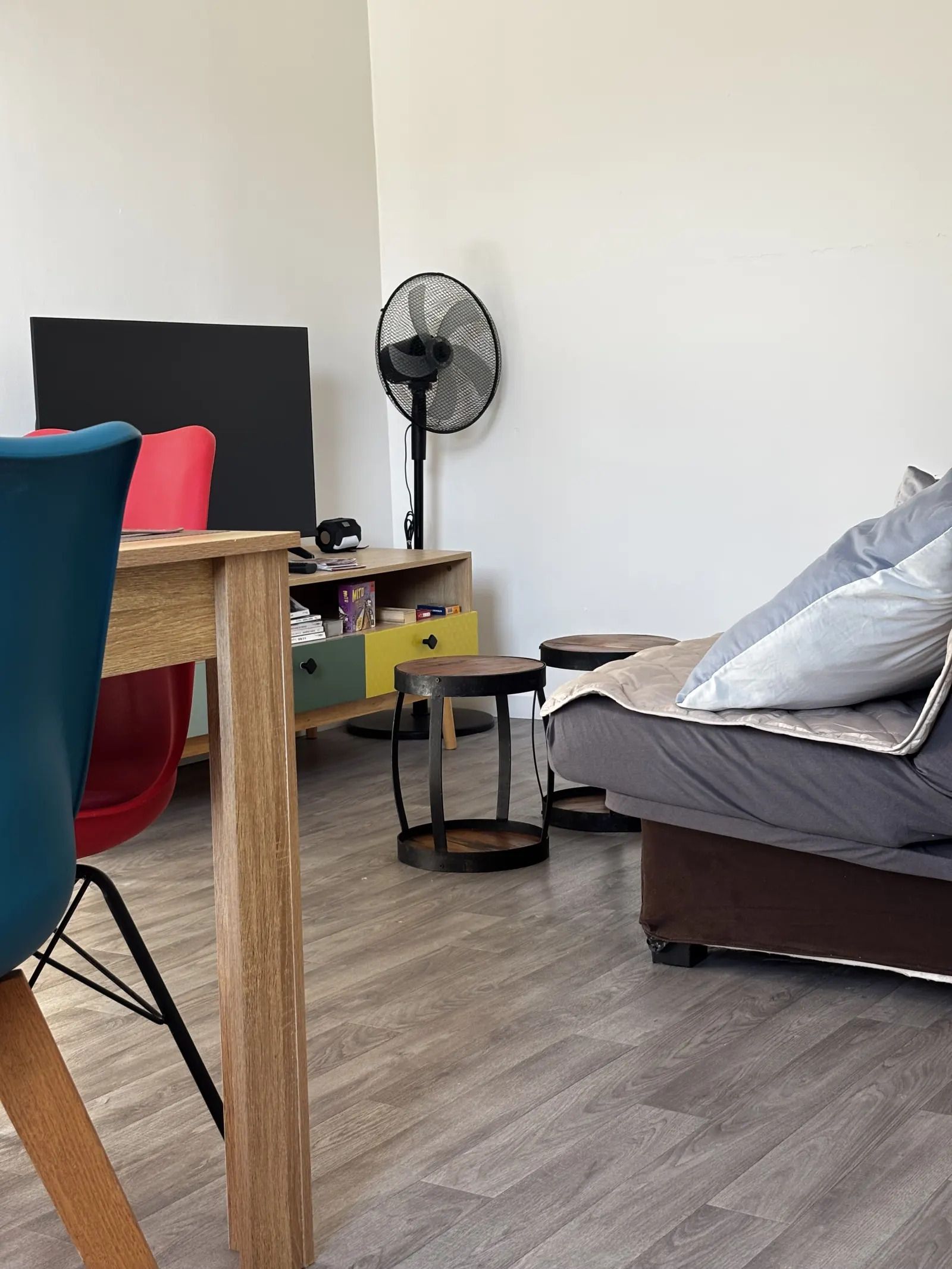 A modern living room with a wooden dining table, a sofa, a small side table, a fan, and a TV.
