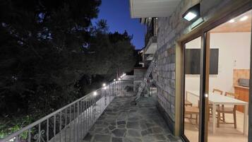 A stone-floored outdoor area with a metal railing, a staircase, and a view of a dining area with tables and chairs.