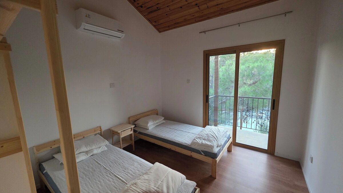 Ein Zimmer mit zwei Holzbett, einem kleinen Nachttisch, einem Fenster mit Blick auf Grünflächen und einer Klimaanlage.