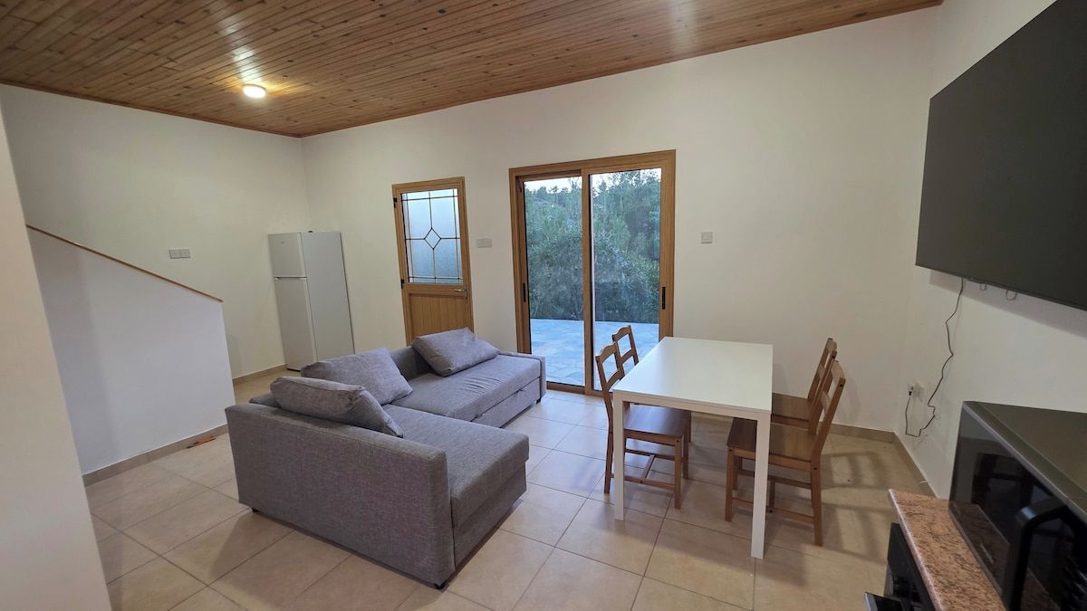 A room with a sofa, a table, chairs, a TV, and a sliding door leading to an outdoor area.