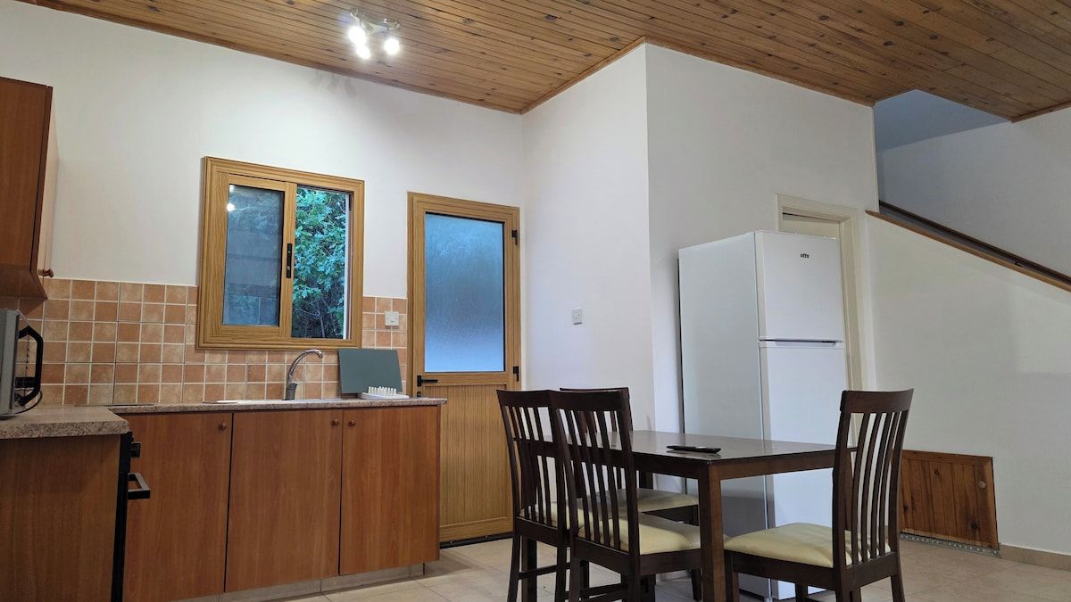 A kitchen with wooden cabinets, a dining table with chairs, a refrigerator, and a window with a view of trees.