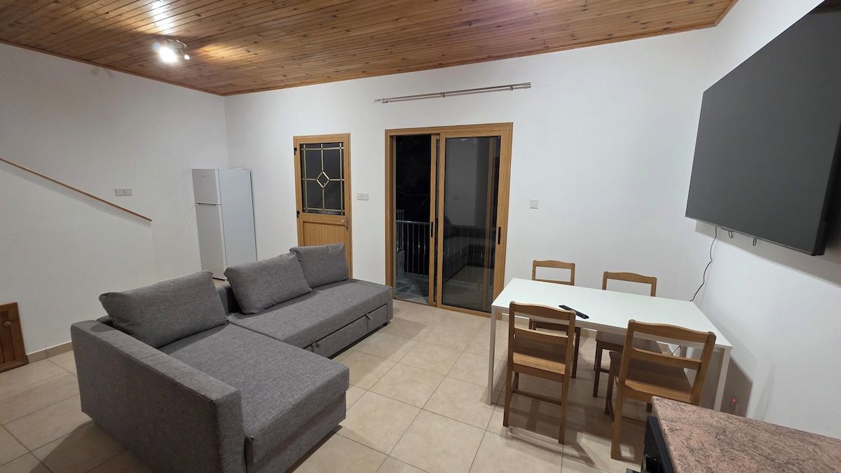 Ein Wohnzimmer mit grauem Sofa, einem Esstisch mit Stühlen, einem an der Wand befestigten Fernseher und einer Glastür zum Außenbereich.