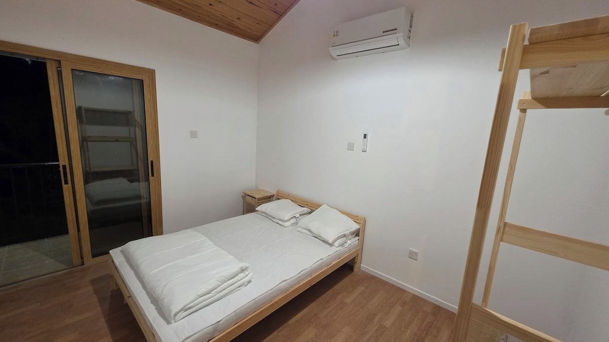 Un lit simple avec un cadre en bois et une literie blanche, dans une pièce dotée d’un plancher en bois et d’un plafond avec des poutres apparentes.