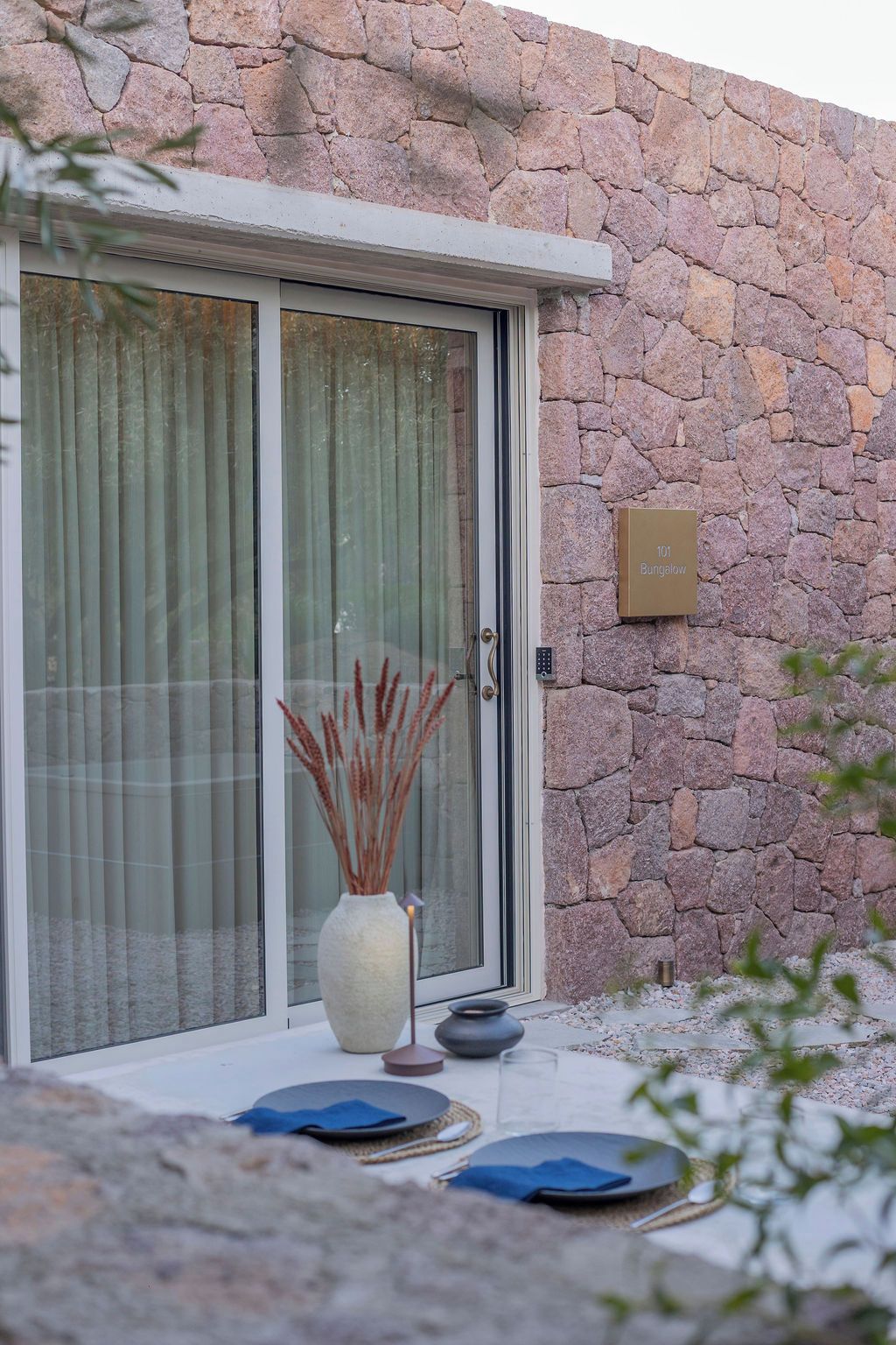 Un muro de piedra con un cristalero corredero, una planta en maceta y un cartel que indica una cabaña.