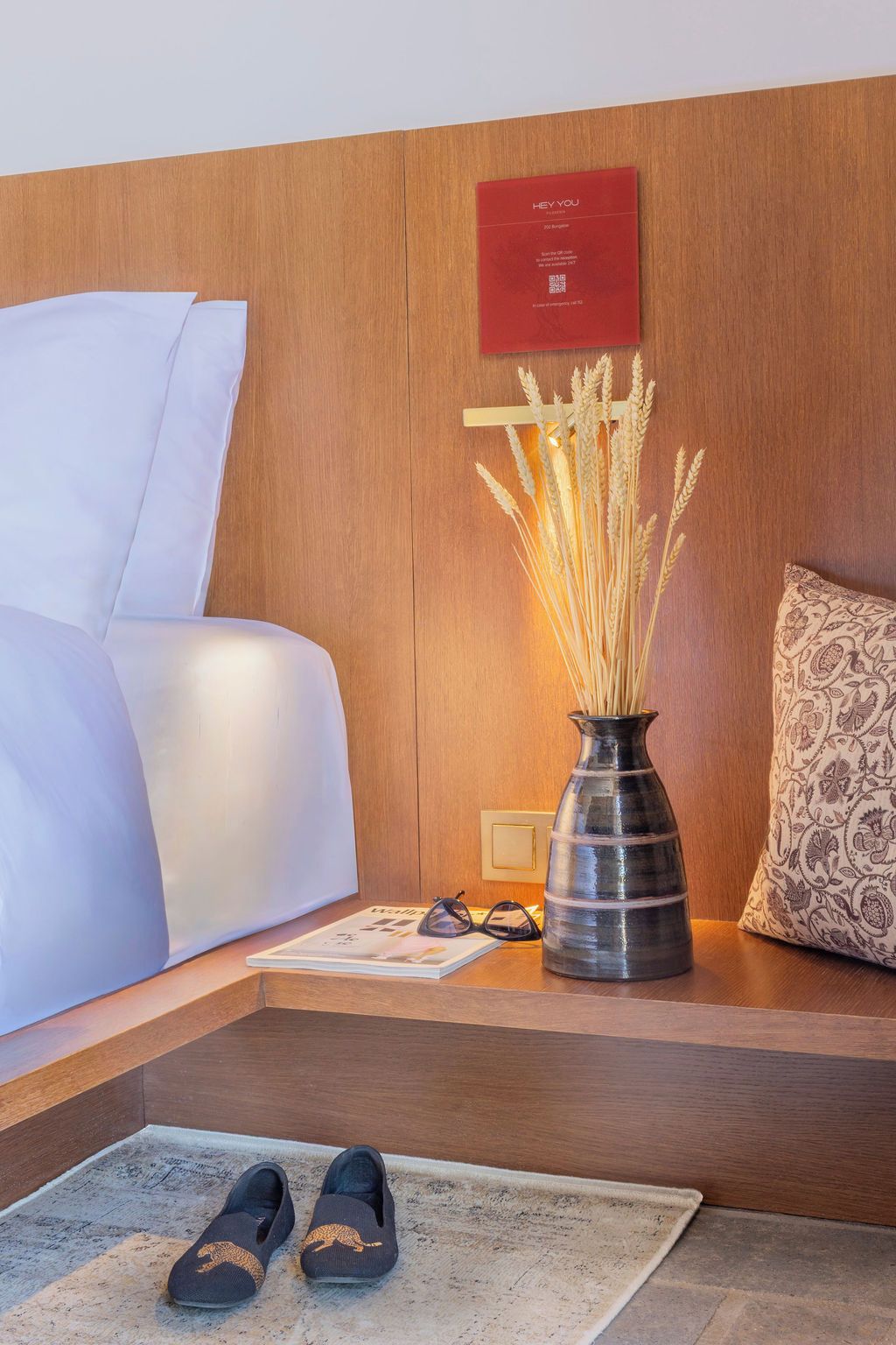 A hotel room with a bed, a bedside table with a vase of wheat stalks, a red plaque, a pair of glasses, and a pair of slippers on the floor.