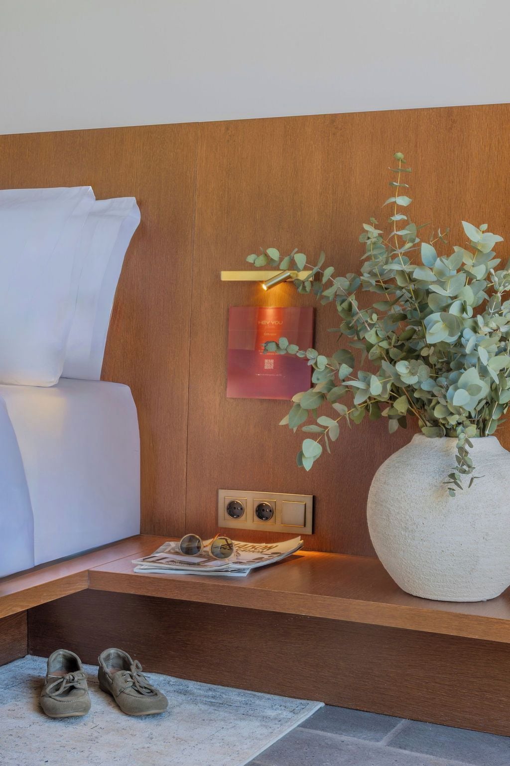 Une chambre d’hôtel bien rangée, avec une tête de lit en bois, une table de chevet ornée d’un vase de plantes, une paire de chaussures et un magazine.