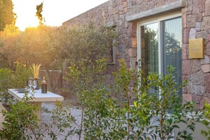 A stone building with a dining area set up outside, a door with a number plaque, and a garden with various plants.