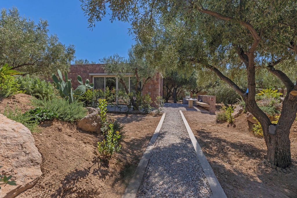 Ein Gartenweg führt zu einem Gebäude mit einer Stein-Fassade, umgeben von verschiedenen Pflanzen und Bäumen.