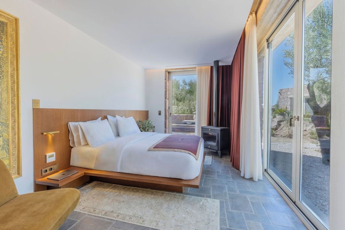 Ein modernes Schlafzimmer mit einem großen Bett, Nachttischen und einer Sitzecke am Fenster mit Blick ins Freie.