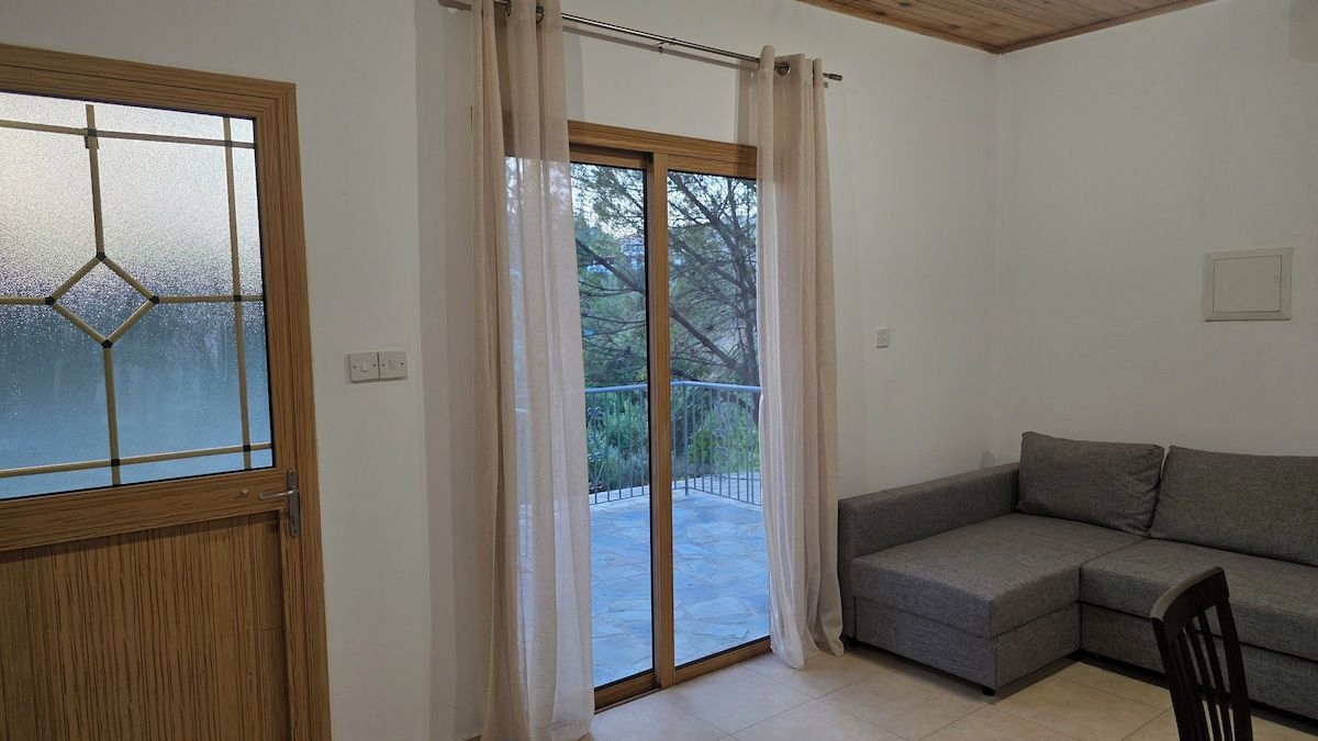 A room with a sliding glass door leading to a balcony, a grey sofa, and a wooden door with a frosted glass window.