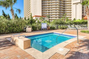 Ein Swimmingpool mit einer gefliesten Kante, umgeben von einer Pflasterterrasse, Palmen und einem mehrstöckigen Gebäude im Hintergrund.