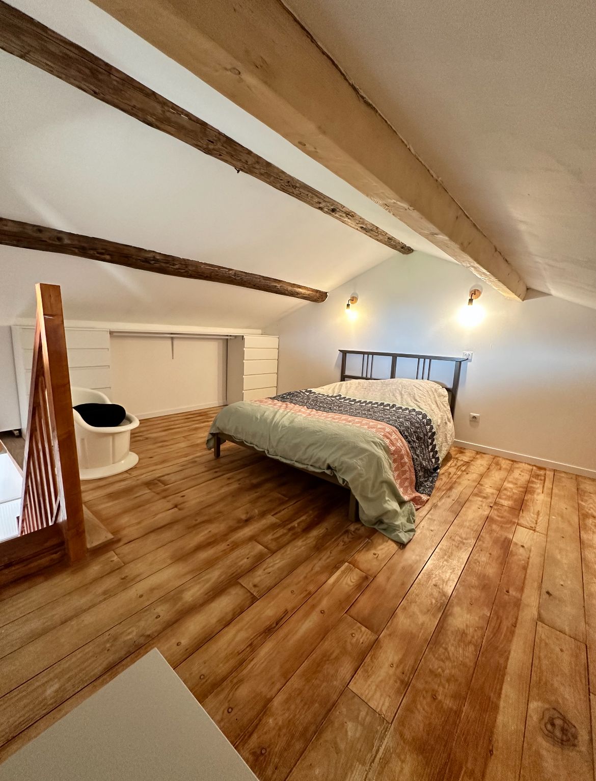 Ein Schlafzimmer mit schrägen Wänden, Holzbalken, einem Bett mit gemustertem Überwurf, einem weißen Stuhl und einem kleinen weißen Schrank.