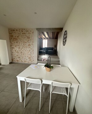 Ein Essbereich mit einem weißen Tisch und Stühlen, einer Topfpflanze und Blick auf ein Wohnzimmer mit einer Couch und einem Fenster.