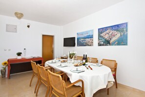 A dining area with a white table set for six, surrounded by wicker chairs. There are two posters on the wall, a telephone, and a small plant on a side table. - Spacious 110 m2 apartment with terrace and garden, just 30m from the beach (Podstrana - Mutogras)