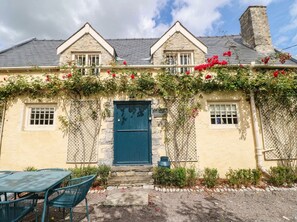 A two-story building with a stone facade, a blue door, and a small garden area with a table and chairs. - The Cheese House at Gileston Manor (Barry)
