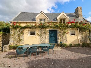 Ein Steinhaus mit einer grünen Tür, einer Schotterterrasse mit grünen Metallstühlen und roten Blumen an der Wand.