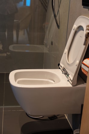 Un inodoro moderno con tapa, un pulsador de descarga visible y un accesorio bidet incorporado en la pared.