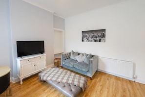 A living room with a flat-screen TV on a white cabinet, a grey sofa with cushions, a plaid throw blanket, and a cityscape photo on the wall. - Cosy Kilmarnock Apartment (East Ayrshire Council)