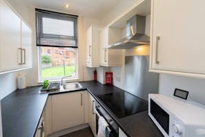 A compact kitchen with a stainless steel range hood, a microwave, and a sink with a window above. - Cosy Kilmarnock Apartment (East Ayrshire Council)