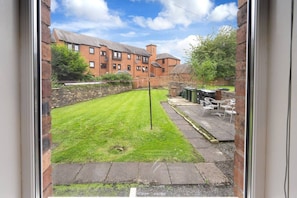 A view of a grassy backyard with a brick wall, a patio with chairs, and a building in the background. - Cosy Kilmarnock Apartment (East Ayrshire Council)