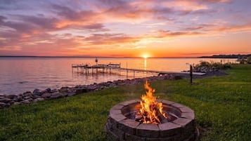 Un foyer extérieur aménagé dans un espace herbeux, offrant une vue sur le coucher de soleil au-dessus d’une étendue d’eau et d’un quai.