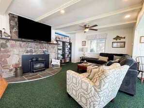 A living room with a stone fireplace, a sofa, a television, and a ceiling fan. - Oneida Lake House | Fire Pit | Dock | Sleeps 9 (Bernhards Bay)