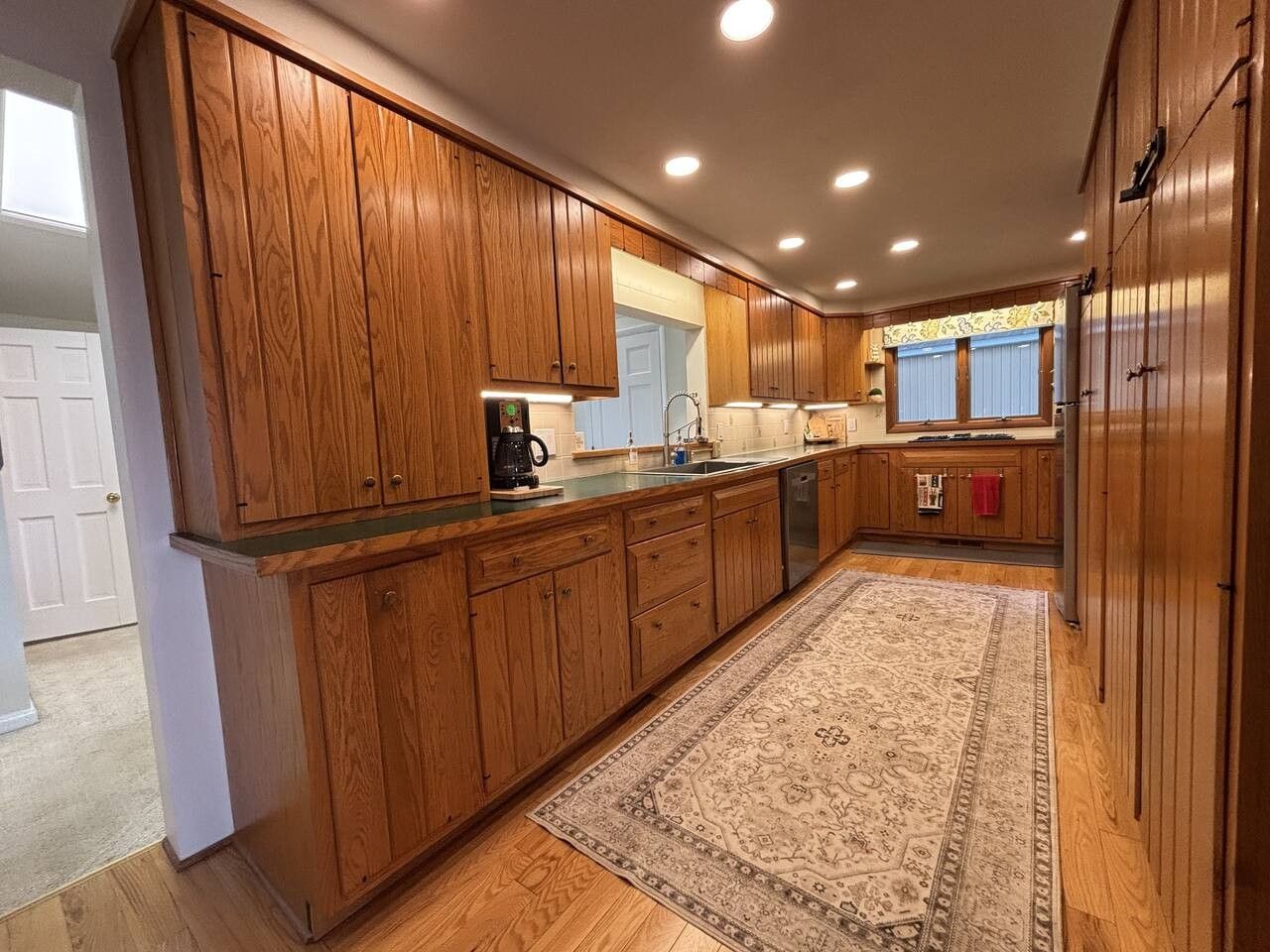 A kitchen with wooden cabinets, a central island, a coffee maker, and a patterned rug.