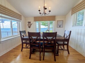 A dining room with a wooden table and chairs, a chandelier, and large windows with a view. - Oneida Lake House | Fire Pit | Dock | Sleeps 9 (Bernhards Bay)