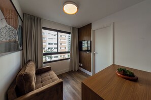 A modern hotel room with a wooden floor, a brown sofa, a wooden table with a potted plant, a television mounted on a wooden panel, and a window with a view of a building. - Agami by Bhomy  (São Paulo)