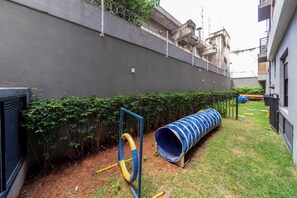 A fenced outdoor area with a blue and yellow play structure, a covered pipe, and a grassy lawn. - Agami by Bhomy  (São Paulo)