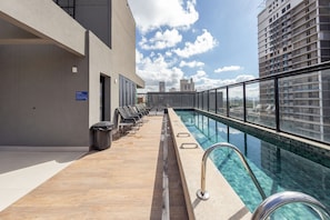 A rooftop pool area with a clear view of the city skyline, a row of lounge chairs, and a trash bin. - Agami by Bhomy  (São Paulo)