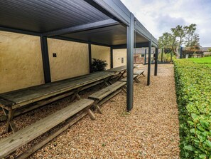 A covered outdoor seating area with wooden benches and tables, gravel ground, and a hedge. - The Carriages (Barry)