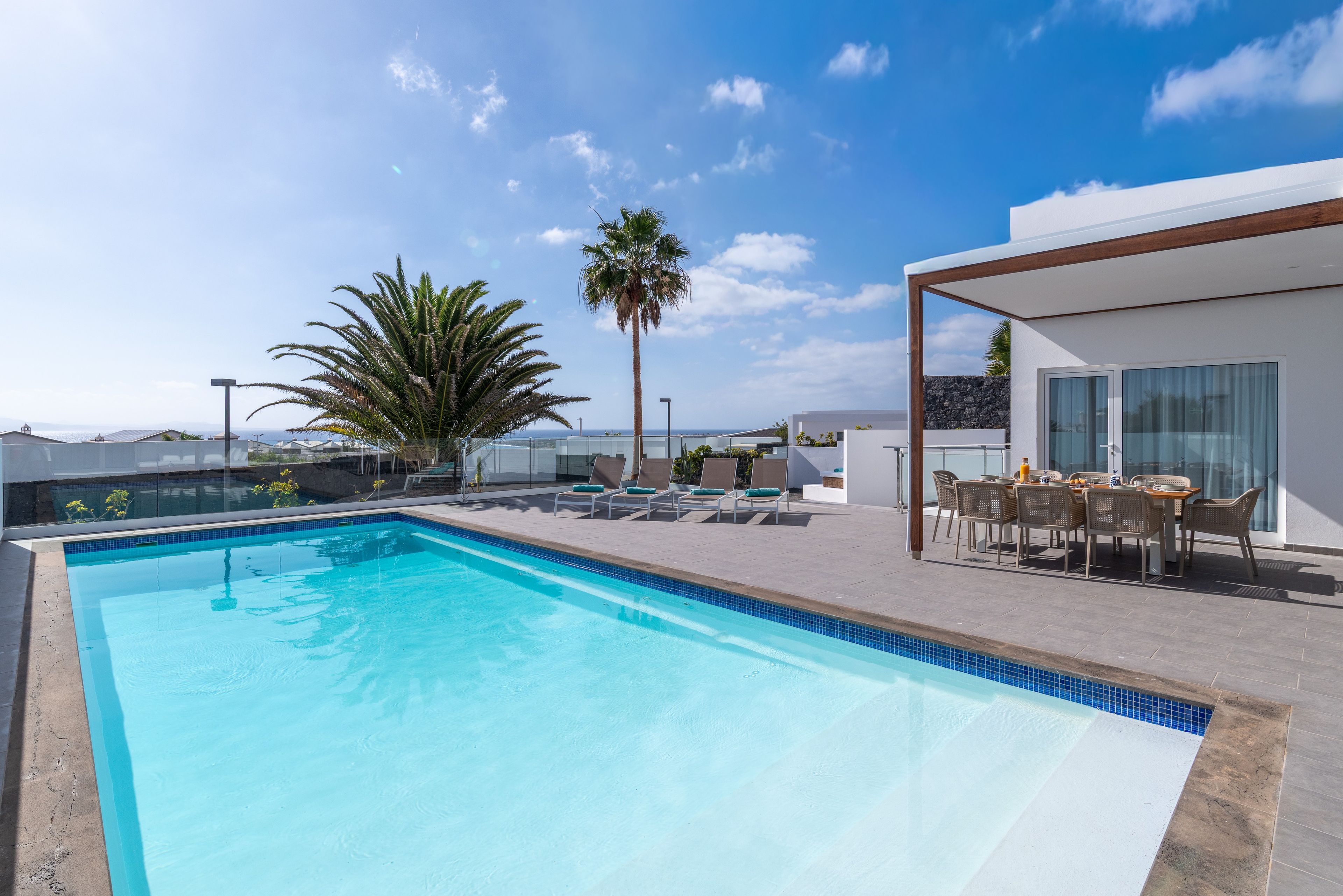 A swimming pool with lounge chairs and a table, surrounded by a modern building with a view of the sea.