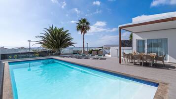 A swimming pool with lounge chairs and a table, surrounded by a modern building with a view of the sea.