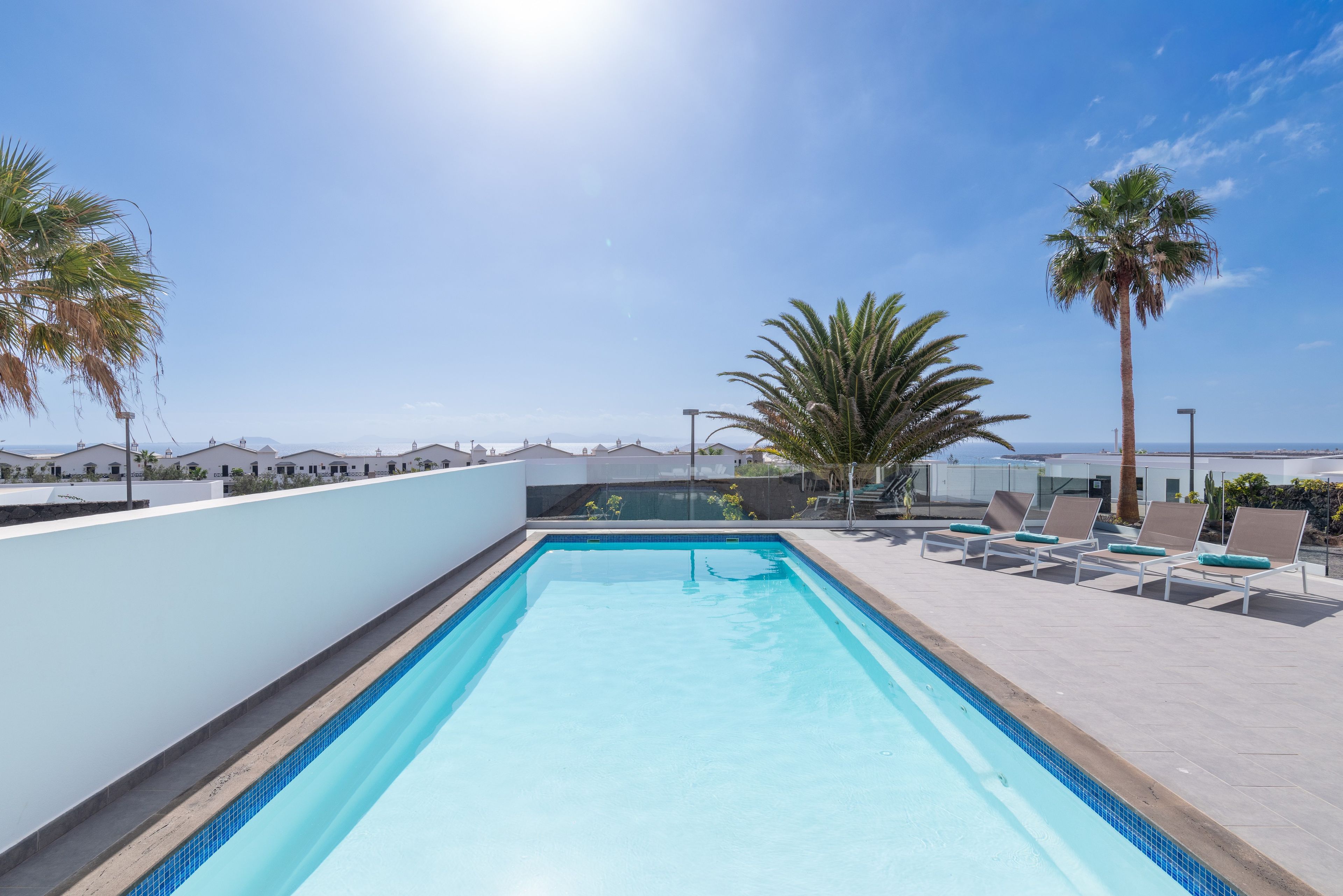 A rooftop swimming pool with clear blue water, surrounded by lounge chairs and palm trees, overlooking a coastal landscape.