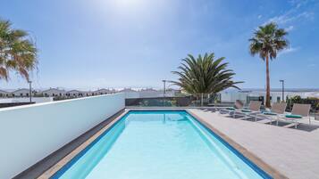 A rooftop swimming pool with clear blue water, surrounded by lounge chairs and palm trees, overlooking a coastal landscape.