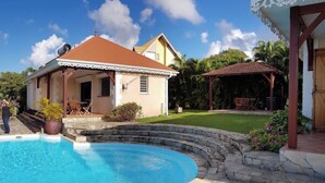 A swimming pool with a stone edge, a house with a red roof, and a covered patio area. - Vrbo Property (Sainte-Luce)