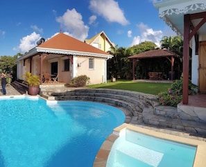 A swimming pool with a stone edge, a house with a red roof, and a covered patio area. - Vrbo Property (Sainte-Luce)