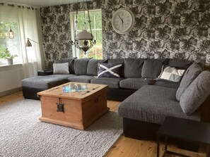 A living room with a sectional sofa, a wooden chest of drawers, a wall clock, and a floral wallpaper pattern. - 4 Star Holiday Home in Munkedal (Munkedal)