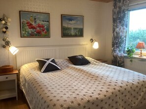 Ein Schlafzimmer mit einem Bett, zwei Wandlampen, einem Nachttisch, einem Fenster mit Vorhängen und zwei gerahmten Bildern.